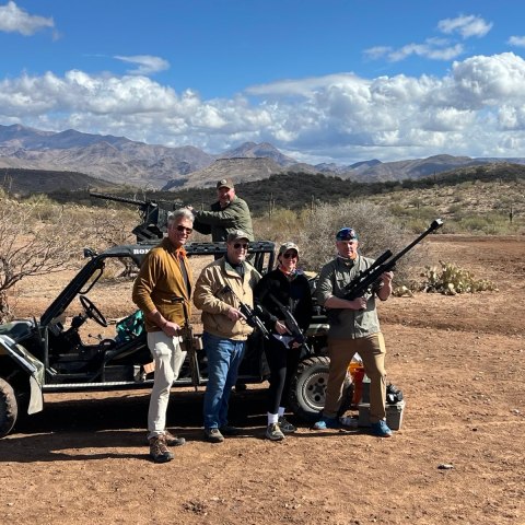 Five people with guns pose by an off-road vehicle in a desert landscape with mountains.