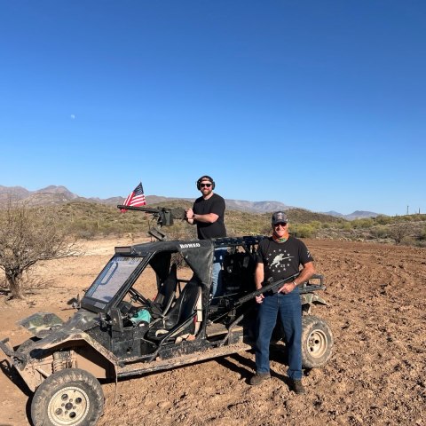 Two people with an off-road vehicle in a desert, one standing on it, holding an American flag.