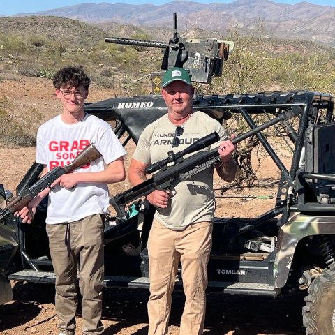 Two people holding rifles stand in front of an off-road vehicle in a desert landscape.