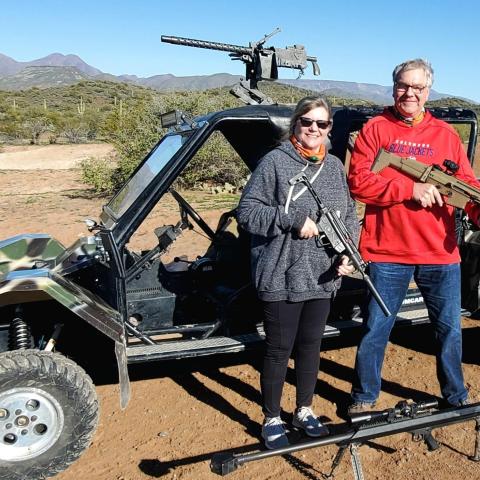 Two people with guns pose in front of an off-road vehicle in a desert landscape.