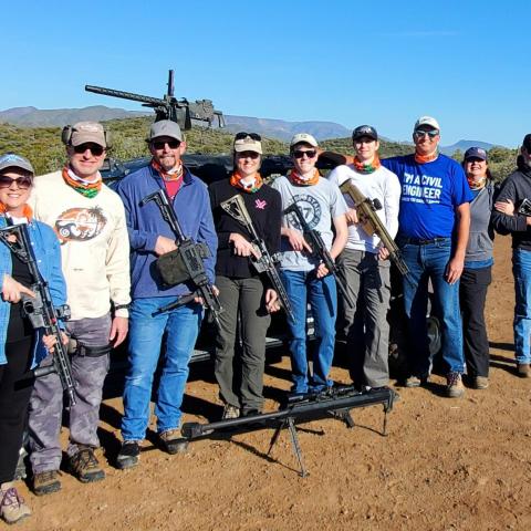 Group of people holding rifles standing in a line outdoors with desert landscape background.