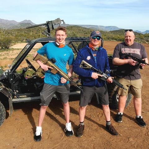 Three men with rifles stand in front of an off-road vehicle in a desert landscape.