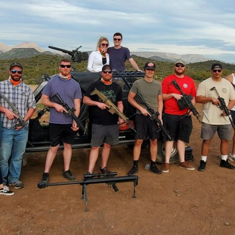 Group of people posing with firearms in front of an off-road vehicle in a desert setting.
