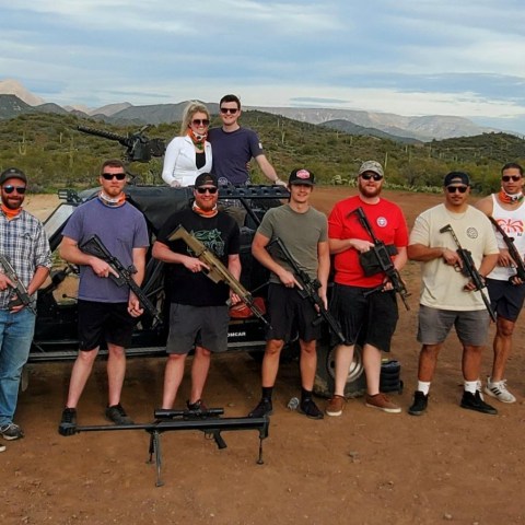 Group of people with rifles standing in front of an off-road vehicle in a desert landscape.