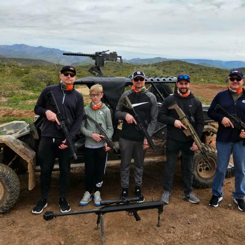Five people holding rifles stand in front of an off-road vehicle in a desert landscape.