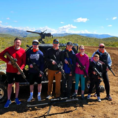 Group of people in outdoor setting holding rifles, with a jeep and mounted gun in the background.