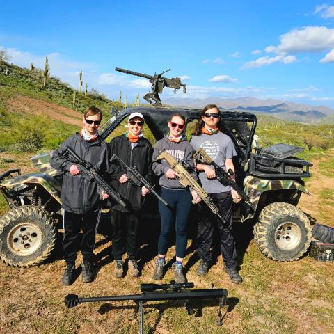 Four people holding guns stand by an off-road vehicle in a desert landscape.