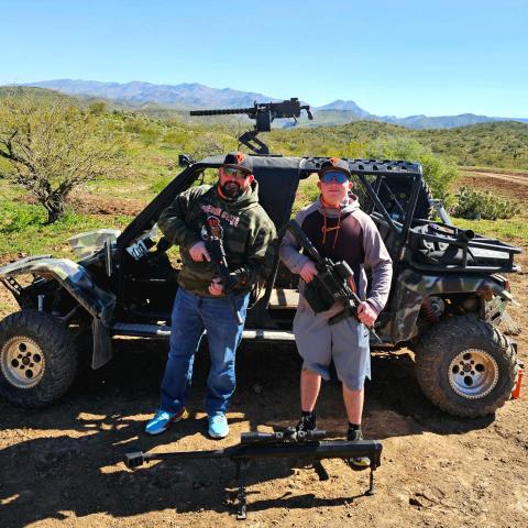 Two men with rifles stand beside an armored off-road vehicle in a desert landscape.