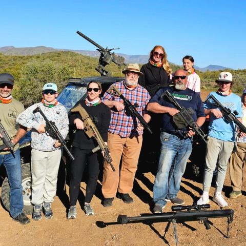 Group of people with firearms pose in front of a military vehicle in a desert landscape.
