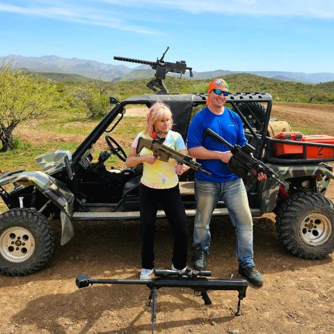 Two people holding guns in front of an off-road vehicle in a desert landscape.
