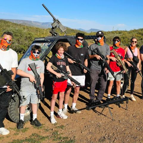 Group of people with masks holding firearms standing in front of an off-road vehicle with a mounted gun.