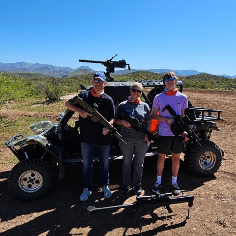 Three people holding rifles in front of an off-road vehicle on a dirt path under a clear blue sky.