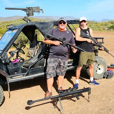 Two men with rifles stand beside an off-road vehicle in a desert landscape.