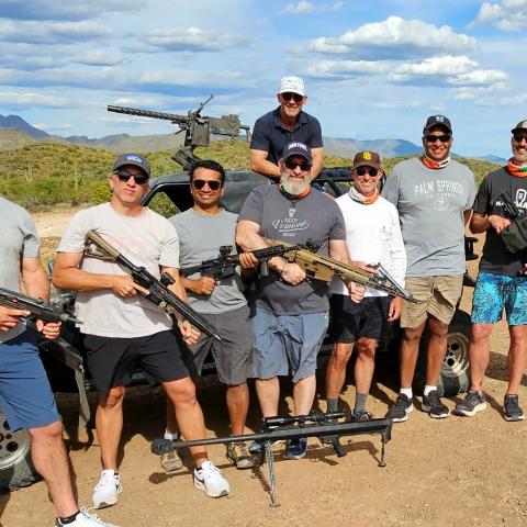 Group of nine men outdoors holding firearms, with a mounted gun in the background.