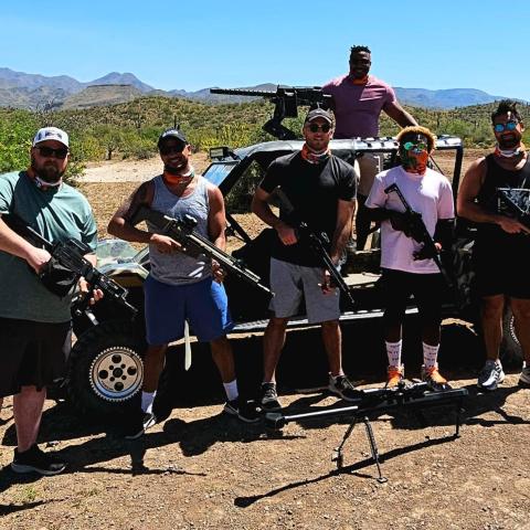 Six men with rifles standing by an off-road vehicle in a desert landscape.