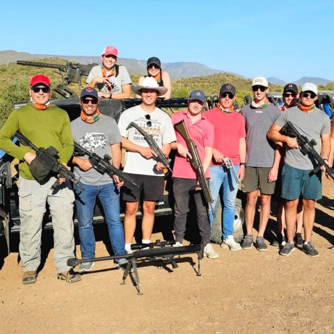 Group of people outdoors holding rifles, posed in front of a vehicle with desert landscape in the background.