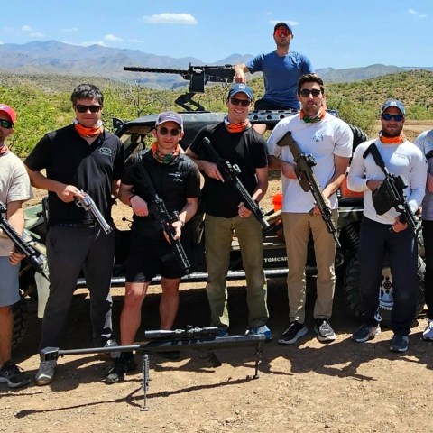 Group of people outdoors holding rifles, standing in front of vehicles with a mountainous background.