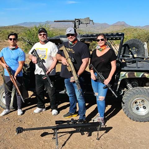 Four people with rifles standing in front of an off-road vehicle in a desert landscape.