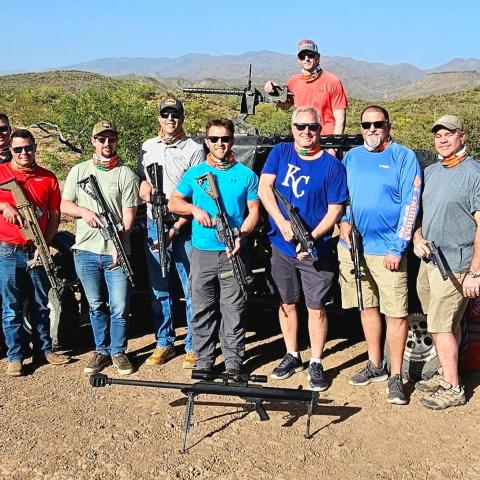 Group of men outdoors holding guns, posing in desert landscape with mountains in background.