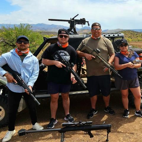 Four people with firearms in front of a vehicle in a desert setting.