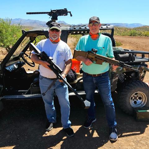 Two men holding rifles in front of an off-road vehicle in a desert setting.