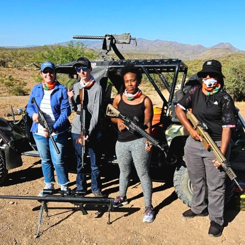Four people holding guns in front of an off-road vehicle in the desert.