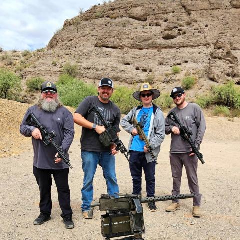 Four men with rifles standing in a rocky desert landscape with a mounted gun in front.
