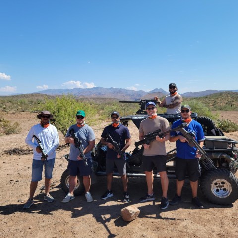 Six people holding rifles in a desert near an ATV with mountains in the background.