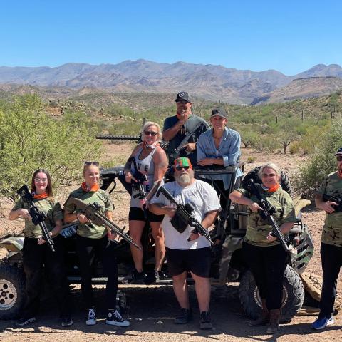 Group of eight people with guns posing outdoors near an off-road vehicle in a desert landscape.
