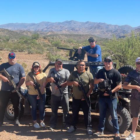 Group of seven people holding rifles, standing by an off-road vehicle in a desert landscape.