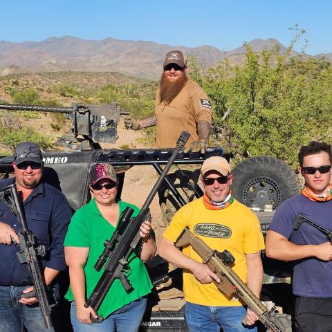 Five people with rifles pose in front of an off-road vehicle in a desert landscape.