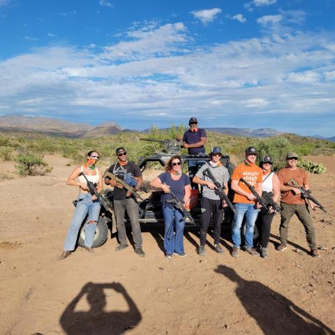 Group of people with guns posing by a vehicle in a desert landscape under a blue sky.