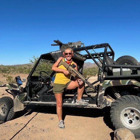 Person standing on an off-road vehicle in desert, holding a rifle, with clear blue sky.