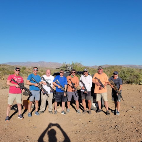 Eight men holding rifles pose in a desert landscape under a clear blue sky.