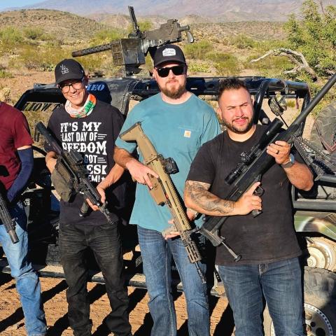 Four men holding rifles stand in front of an off-road vehicle in a desert setting.
