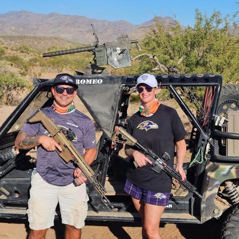 Two people with rifles stand by an off-road vehicle in a desert landscape.