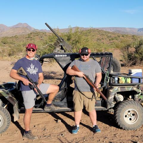 Two men with rifles stand beside a small off-road vehicle in a desert landscape.