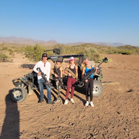 Four people with rifles stand by an off-road vehicle in a desert landscape.