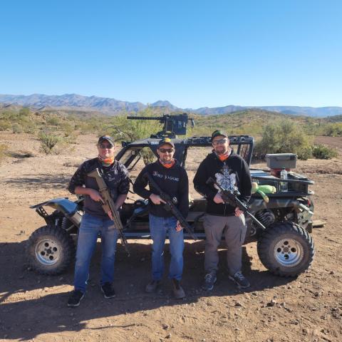 Three people with rifles stand in front of an off-road vehicle in a desert landscape.
