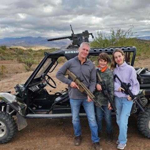 Three people holding rifles stand beside a dune buggy in a desert landscape.