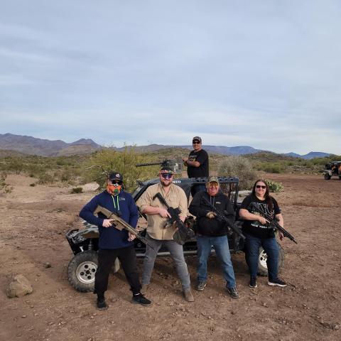 Five people with rifles pose in front of an off-road vehicle in a desert landscape.
