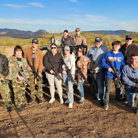 Group of people holding rifles, standing outdoors on desert terrain with a mountain in the background.
