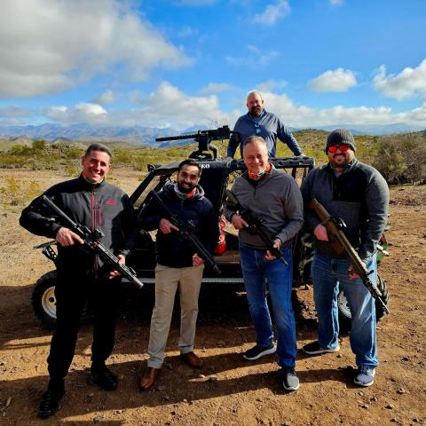 Five people with rifles stand outdoors in front of a rugged vehicle on a sunny day.