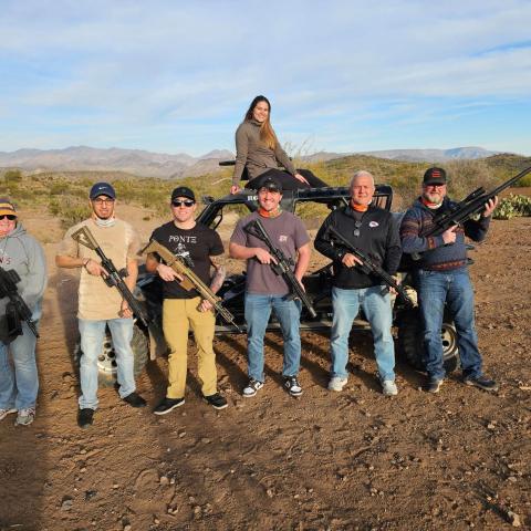 Group of seven people holding rifles, standing in desert landscape near a vehicle.