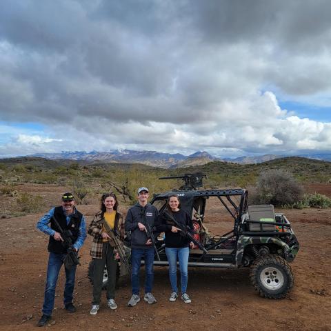 Four people with rifles stand next to an off-road vehicle in a desert landscape under a cloudy sky.