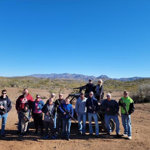 Group of people posing with rifles in a desert landscape with mountains and clear blue sky.