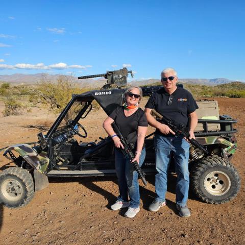 Two people holding rifles stand by a dune buggy in a desert landscape.