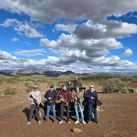Group of six men with rifles pose in desert terrain with cloudy sky backdrop.