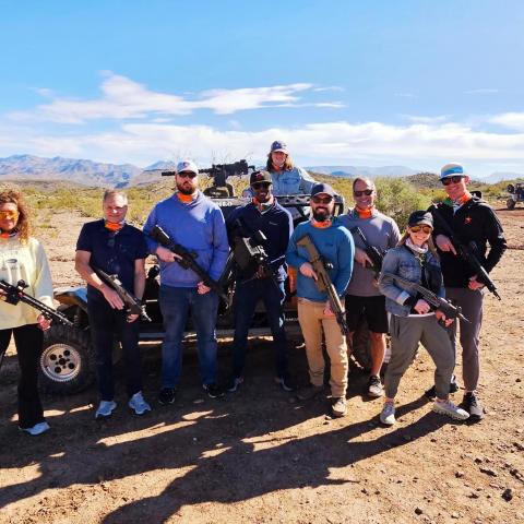 Group of nine people with rifles standing outdoors in a desert landscape.