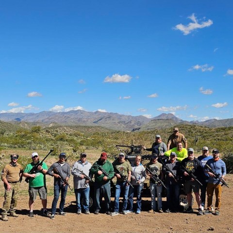 Group of people with rifles outdoors in a desert landscape under a clear blue sky.